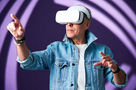 Senior Man In Casualwear And Vr Headset Pointing At Something While Traveling In Futuristic Reality Against Lavender Background