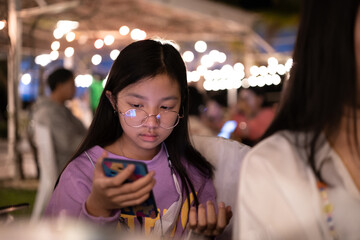 Asian girl in eyeglasses is busy looking at smart phone in restaurant at night.