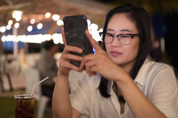 Asian woman in eyeglasses is busy looking at smart phone in restaurant at night.