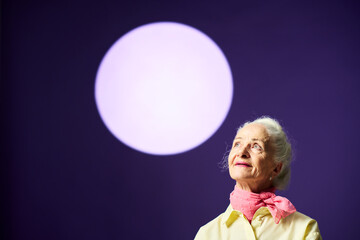 Stylish aged woman with white hair looking upwards at full moon on violet sky while standing in front of camera during photo session in studio