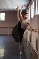 Graceful Asian ballerina in a beige bodysuit and black skirt is rehearsing in a dance class. 