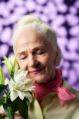 Happy senior woman with grey hair enjoying nice smell of gorgeous white lily while standing in front of camera during photo session