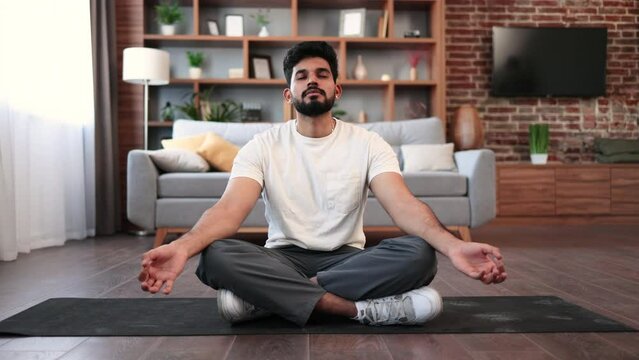 Relaxed indian man in sportswear practicing meditation in lotus pose at cozy living room. Young bearded guy keeping hands in mudra gesture and breathing calmly during workout.