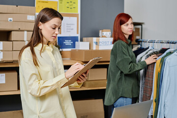 Obraz premium Young brunette woman with document looking through list of people needing new warm clothes against her colleague standing by rack