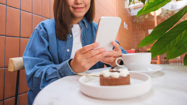 Closeup image of a young woman holding and using mobile phone with cake and coffee cup on the table in cafe