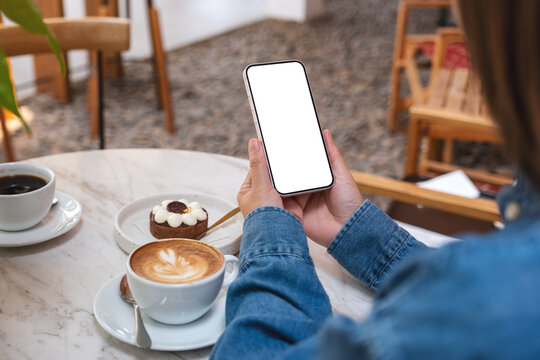 Mockup image of a woman holding and using mobile phone with blank desktop screen with cake and coffee cups on the table in cafe