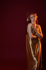 Close up fantasy portrait of beautiful woman model with red hair, goddess silk robes & gold crown.  Posing with gestural hands reaching out, isolated on dark red studio background 