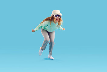 Fototapeta na wymiar Studio shot of funny crazy screaming girl in fashionable outfit. Young woman in hat and sunglasses feeling happy and excited or furious and enraged screaming loudly isolated on blue background