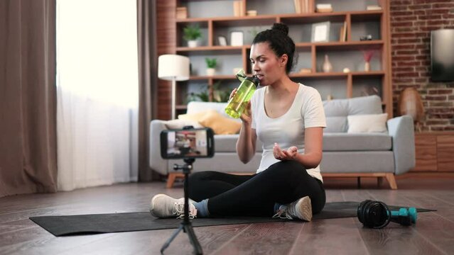 Focused african american female in gym clothes drinking water from sports bottle while sitting cross-legged in front of active cell phone on tripod. Fit sports vlogger showing workout routine at home.