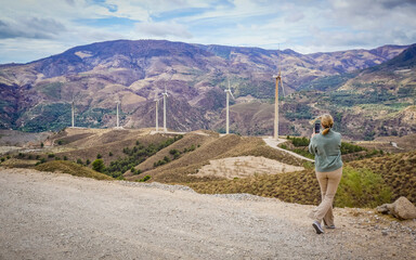 Fototapeta na wymiar Blond young woman taking photos of a mountain landscape with wind turbines