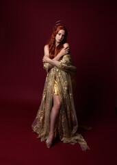 Fototapeta na wymiar Full length fantasy portrait of beautiful woman model with red hair, goddess silk robes & gold crown. Standing pose gestural hands reaching out isolated on dark red studio background 