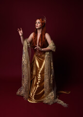 Obraz na płótnie Canvas Full length fantasy portrait of beautiful woman model with red hair, goddess silk robes & gold crown. Standing pose gestural hands reaching out isolated on dark red studio background 
