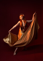 Full length fantasy portrait of beautiful woman model with red hair, goddess silk robes & gold crown. Standing pose gestural hands reaching out isolated on dark red studio background 