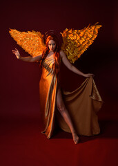Full length portrait of beautiful woman model with long red hair, gold silk robes, crown & fantasy feather angel wings. Standing pose gestural hands reaching out isolated on dark red studio background