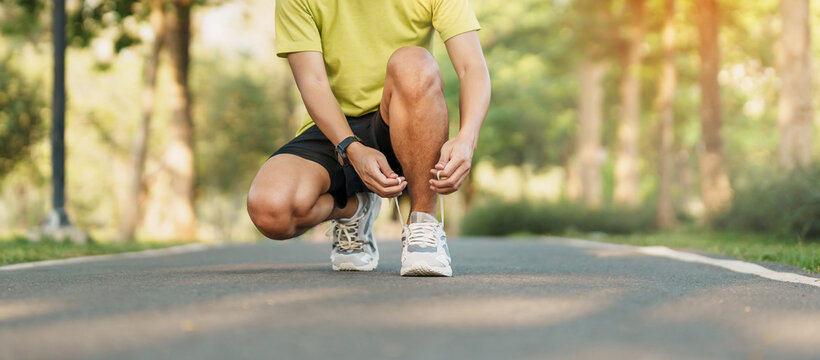 Young athlete man tying running shoes in the park outdoor, male runner ready for jogging on the road outside, asian Fitness walking and exercise on footpath in morning. wellness and sport concepts