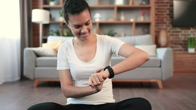 Young multiracial female looking at activity fitness tracker while relaxing on floor in apartment. Cheerful fit woman checking heart rate on modern smartwatch while having break from home workout.