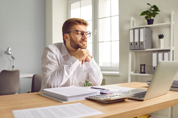 Young financial accountant or business man at his office desk. Happy, satisfied man in a white shirt sitting at his working table with an accounting book and looking at his modern laptop computer