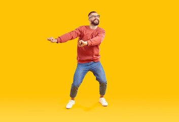 Fototapeta na wymiar Cheerful, playful, funny and positive bearded man in glasses, red sweatshirt and jeans in good mood having fun dancing on yellow background, full length portrait. Banner for advertisement, marketing.