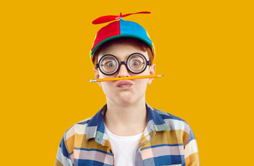Studio portrait of funny preteen boy who is fooling around holding pencil as mustache. Child boy in funny glasses and cap holding balancing pencil between lips and nose isolated on orange background. - obrazy, fototapety, plakaty