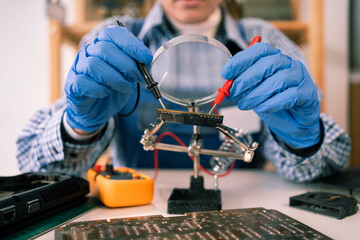 Engineer testing microcircuit through magnifier using multimeter. Electronic renovation in repair shop. Female repairman tinkering computer component, close-up.