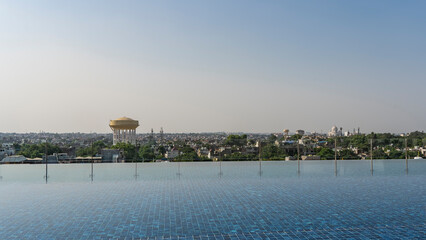 Naklejka premium Infinity outdoor swimming pool on the roof. The tiled blue bottom is visible through the water. A transparent barrier along the edge. In the distance - a panorama of the city against the sky. India