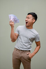 Shocked and excited Asian man holding USD dollar bills, standing against a green studio background