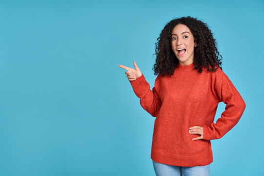 Young excited surprised latin woman pointing finger aside isolated on blue background. Amazed female model showing advertising great choice presenting sale promotion shopping offer concept.
