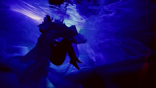 In slow motion, the silhouette of an elegant girl under water, as in a fairy-tale dream, floats in weightlessness in neon lights