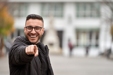 Caucasian man with glasses and black coat smiling and pointing to camera in a city square.
