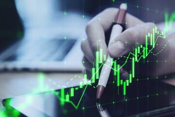 Close up of hand using digital tablet with pen and growing green candlestick chart on blurry desktop background. Forex, trade and finance concept. Double exposure.