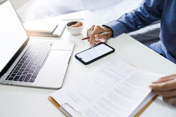 Fototapeta premium Businessman using smart phnoe and reviewing document reports at office workplace with computer laptop. legal expert, professional lawyer reading and checking financial documents or insurance contract