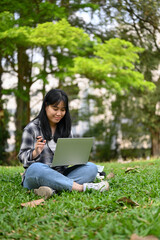 Pretty Asian female college student using her laptop and sitting on grass in the campus park.