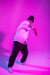 Handsome professional breakdancer man in fashion outfit with hat, white t-shirt, pants and sneakers dances in creative studio with pink and neon light