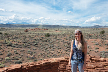 Naklejka premium Portrait of a smiling long-haired blonde young girl with backwards cap in the arid desert in summer, she carries her backpack and photo camera