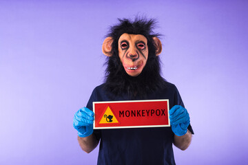 Disguised person with a monkey mask and a doctor's suit, holding a sign with the danger sign and a monkey, which reads: MONKEYPOX, on a purple background. 