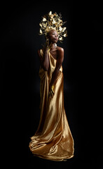  fantasy portrait of beautiful african woman model with afro, goddess silk robes and ornate floral wreath crown. gestural Posing holding golden flowers. isolated on dark  studio background 