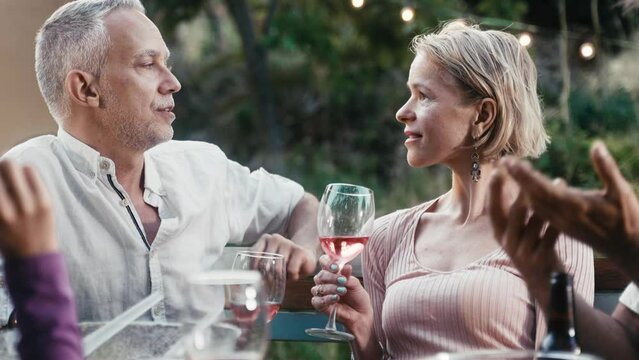 Adult caucasian couple enjoying romantic conversation at garden party on a summer evening, drinking wine, having fun talking with friends