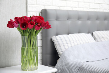 Fototapeta na wymiar Bouquet of beautiful tulips in glass vase on white nightstand near bed