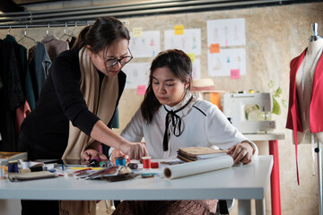 Obraz premium Asian middle-aged female fashion designer teaches a young teen trainee tailor in studio with colorful thread and sewing fabric for dress design collection ideas, professional boutique small business.
