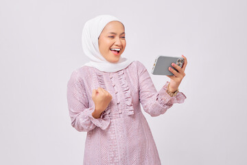 Excited beautiful young Asian Muslim woman wearing hijab and purple dress using smartphone for playing mobile game, celebrating success isolated on white studio background