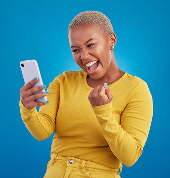 Black woman, phone and fist celebration in studio, blue background and winning online prize. Happy female model celebrate mobile promotion, bonus and excited for deal, success and competition winner