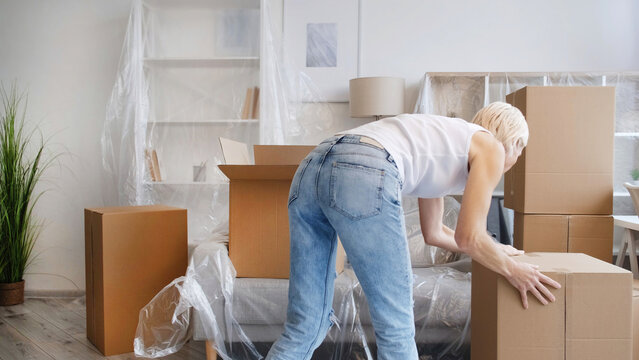 Moving day. Home relocation. Package delivery. Unrecognizable woman carrying carton boxes in light modern apartment with covered furniture.