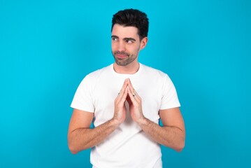 Charming cheerful Young man wearing white T-shirt over blue studio background making up plan in mind holding hands together, setting up an idea.