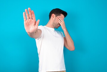 Young man wearing white T-shirt over blue studio background covers eyes with palm and doing stop gesture, tries to hide from everybody.