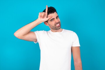 Young man wearing white T-shirt over blue studio background making fun of people with fingers on forehead doing loser gesture mocking and insulting.