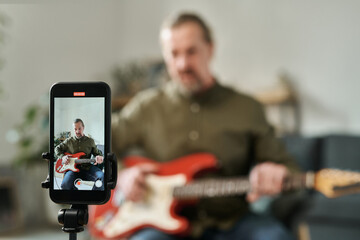 Selective focus of screen of smartphone with guitarist recording online lessons on it at home