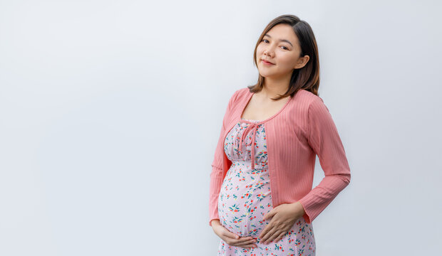 Happy young pragnent Asian woman in maternity clothes smiling and standing in front of a white background