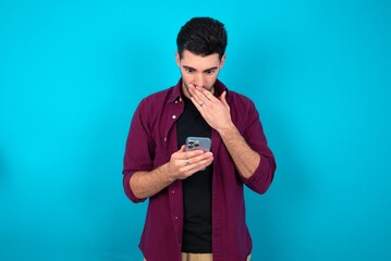 Young man standing over blue studio background being deeply surprised, stares at smartphone...
