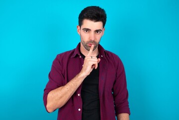 Surprised Young man standing over blue studio background makes silence gesture, keeps finger over lips and looks mysterious at camera