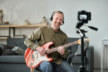 Happy mature man using camera to play guitar online for his musical blog